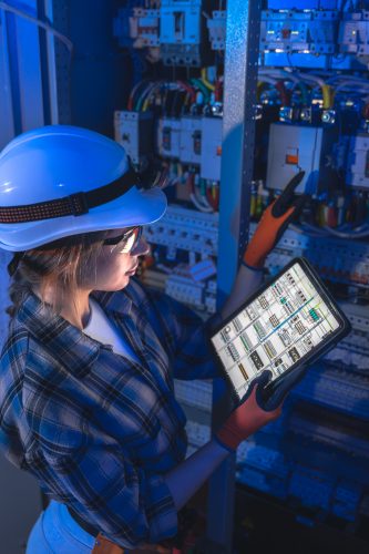 Wider working view in blue light, a specialist balances cabinet control with digital documentation on a tablet. Reliability driven scene focused on uptime, predictive diagnostics, and smart operations.